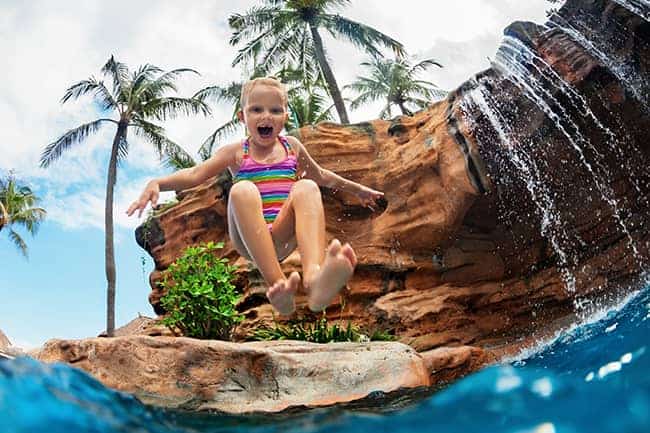 A girl jumping into a pool from a pool jumping rock. 